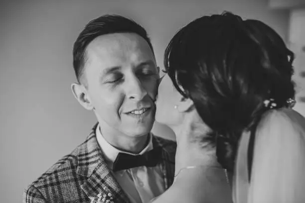 Photo of First meeting is newlywed. Bride kisses groom on cheek, surprise at home. Handsome man embraces woman in room. Groom hugging bride. Happy wedding day of marriage. Closeup couple. Black and white photo