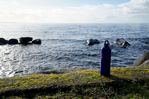 A blue water bottle standing in front of a beautiful and calm lake during a sunny day.