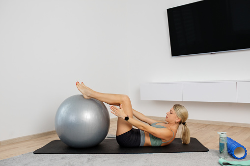 Millennial woman exercising with fitness ball on exercise mat in living room. Fitness, sport and healthy lifestyle concept.