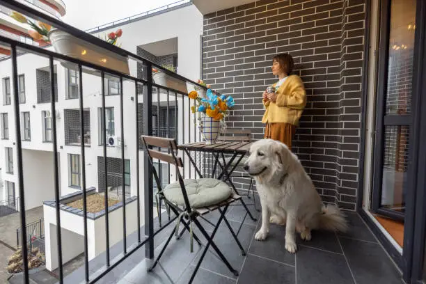 Young woman stands with her dog on balcony, enjoying coffee drink at her property during morning time. City life in a modern residential complex