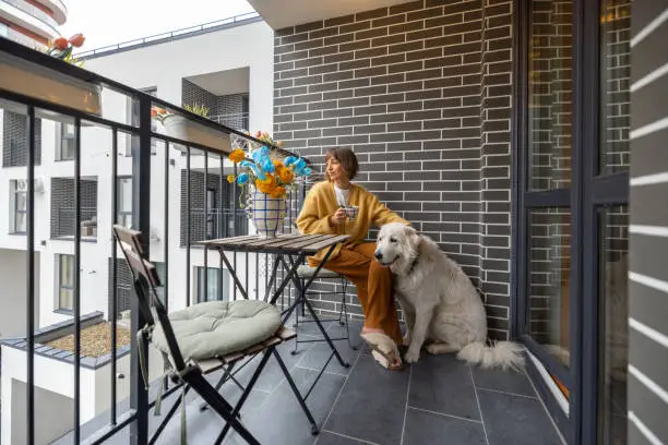 Young woman sits with her dog on balcony, enjoying coffee drink at her property during morning time. City life in a modern residential complex