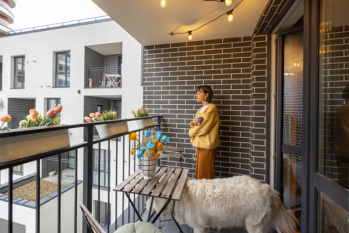 Young woman stands with her dog on balcony, enjoying coffee drink at her property during morning time. City life in a modern residential complex