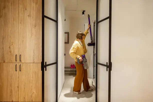 Woman takes vacuum cleaner from wardrobe in hallway at her modern apartment. View through the arch door. Domestic routine and house chores concept