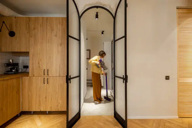 Woman takes vacuum cleaner from wardrobe in hallway at her modern apartment. View through the arch door. Domestic routine and house chores concept