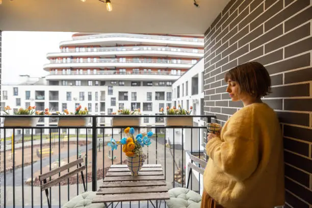 Young woman enjoying coffee drink on balcony of her modern property during morning time. City life in a modern residential complex