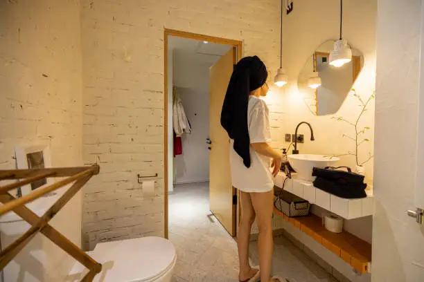 Woman with towel on hair takes care of herself after a shower, looking on mirror in stylish bathroom interior. Wellness and domestic lifestyle concept