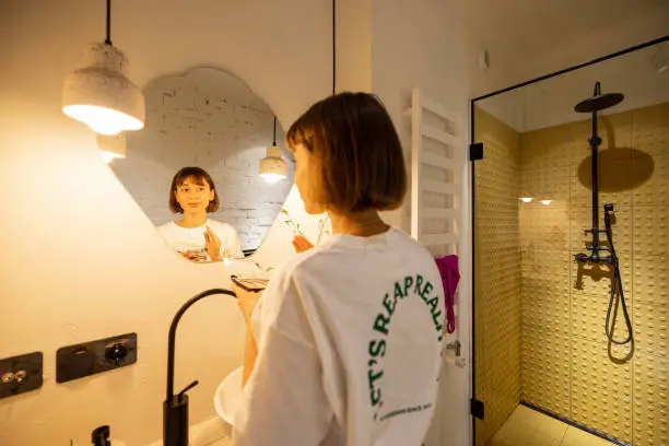 Woman takes care of herself, looking on mirror in stylish bathroom interior. Wellness and domestic lifestyle concept