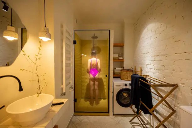 Woman in pink swimsuit takes a shower in the shower cabin at home. Wide interior view of stylish white bathroom. Concept of everyday routine and domestic lifestyle