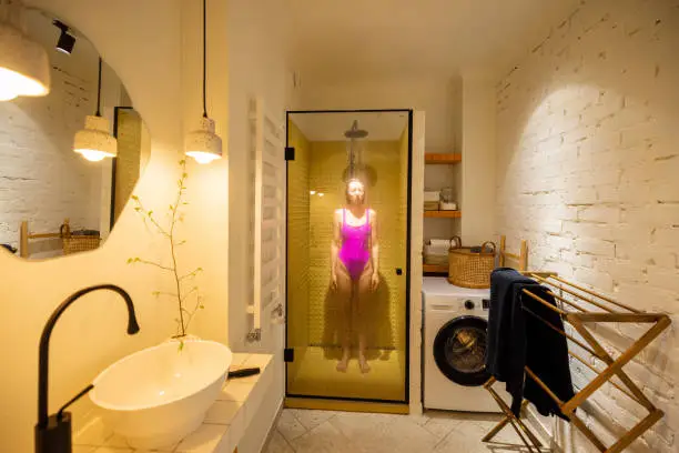 Woman in pink swimsuit takes a shower in the shower cabin at home. Wide interior view of stylish white bathroom. Concept of everyday routine and domestic lifestyle