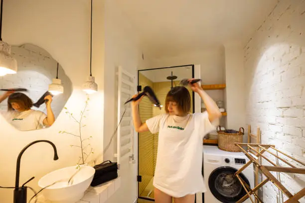 Young woman drying hair with hairdryer after a shower in white and stylish bathroom. Person slightly is motion blurred. Wellness and domestic lifestyle concept