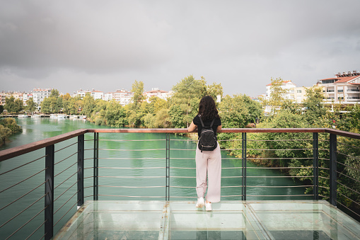 Young woman with a backpack on her back stands on the Manavgat bridge and looks at the Manavgat city