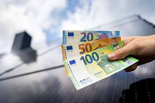 Euro banknotes held by a hand. The background shows the roof of a house covered by solar panels.