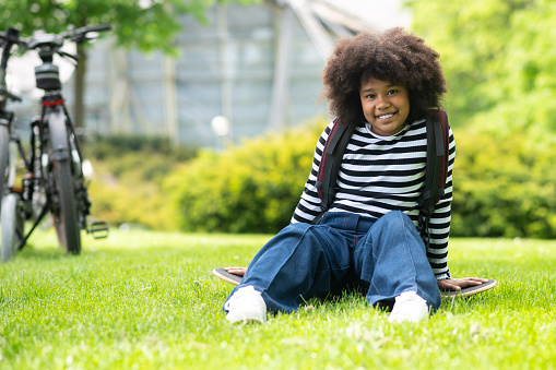 Happy little african american girl wearing striped shirt and jeans sitting on grass in park on summer.
