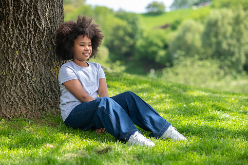 Dreaming little girl sitting on grass under tree wearing white t-shirt and jeans resting in nature enjoying freshair.