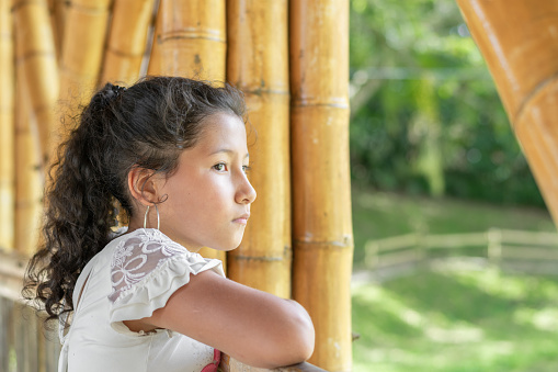 Little Latina girl with brown skin, very serious and bored, thinking about the bullying she receives, standing on the bamboo bridge.