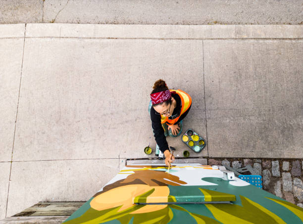 Young Latin woman artist painting sidewalk box mural Young Latin woman artist painting sidewalk public utility box  mural .  She is dressed in casual work clothes. Exterior of public sidewalk in downtown Toronto, Canada. Drone point of view. street art mural stock pictures, royalty-free photos & images