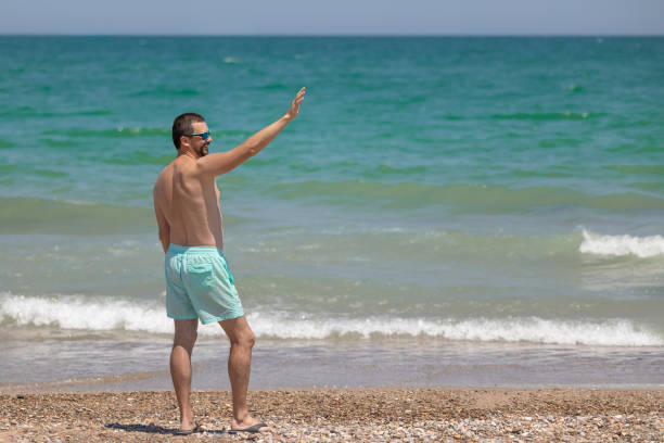 A young white man in sunglasses with a beard stands on the shores of the Mediterranean Sea and waves his hand.