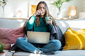 Cold woman working on laptop while drinking hot tea sitting on couch at home