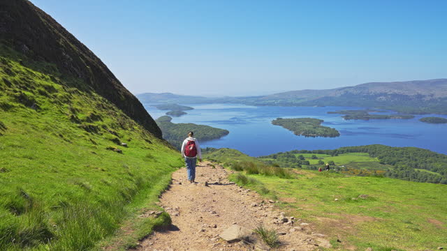 Parallax movement of a trekker walking on a descending path surrounded by a grassy hillside meadow with views of Loch Lomond from the West highland Way, Scotland