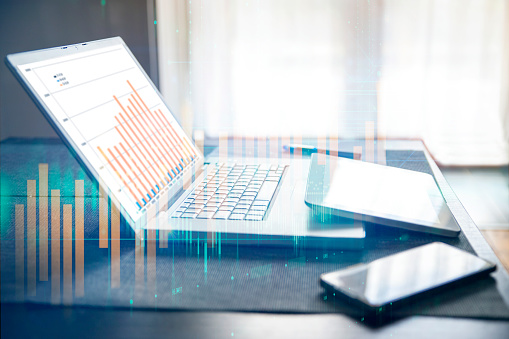 Laptop and digital tablet on work desk with Stock market investment graph and financial numbers.  Abstract background graphic overlay. Close up