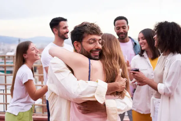 Female and male friends hugging surrounded by a group of people gathered outside in a rooftop. Two people embraced outdoors during a meeting. Greeting at a party.