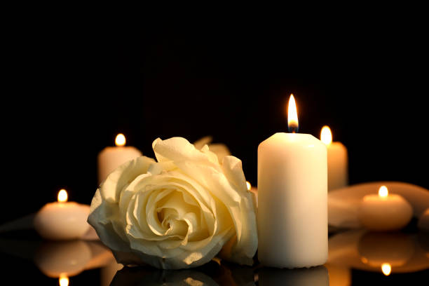 暗闇の中で黒い鏡の表面に白いバラと燃えるろうそく、テキスト用のスペースを持つ接写。葬儀のシンボル