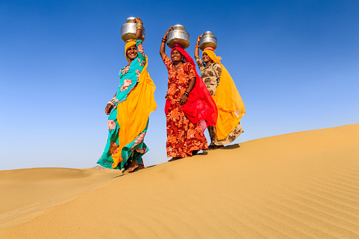 Indian women crossing sand dunes and carrying on their heads water from local well, Thar Desert, Rajasthan, India. Rajasthani women and children often walk long distances through the desert to bring back jugs of water that they carry on their heads.