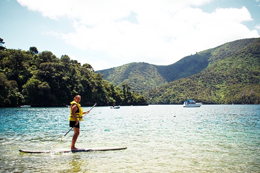 A woman stands whilst paddling on a paddle board. Taken in the coastal waters of the Marlborough Sounds, New Zealand.