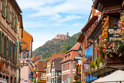 A picturesque, colorful streets in the medieval village of Ribeauville, in the Alsace wine region with Castle Saint-Ulrich in view on the hill behind.