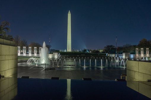 Nighttime panoramic view of the Washington monument and the World War II Memorial located on the National Mall in Washington DC. The Washington monument (c. 1884) is the tallest obelisk in the world. The World War II memorial (c. 2004) features pillars, triumphal arches and fountains honoring veterans of WWII.
