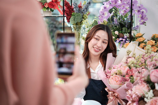 E-commerce business, young Asian female florist worker demonstrates and shows floral arrangements via online live streaming by smartphone application in bright flower shop, beautiful blossoms store.