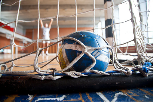 Handball ball in goal with male player celebrating. Low angle, ball in focus, unrecognizable Caucasian person in background.