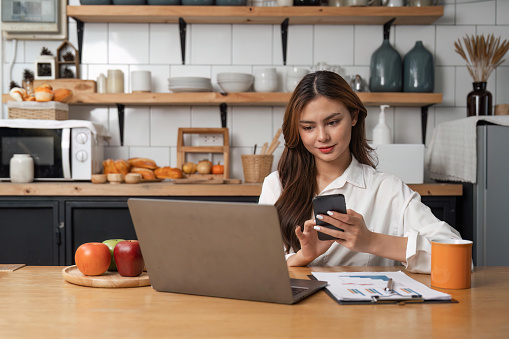 Young woman use cellphone sit at table in kitchen share text messages in social media, enjoy new , watch internet content take break from work on laptop. mobile app usage concept.