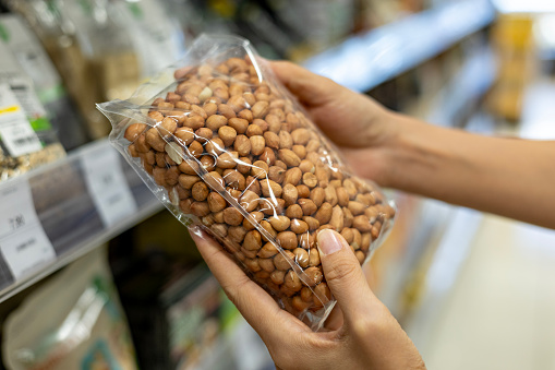 Young Asian woman doing grocery shopping in a supermarket, close up of her hand choosing a pack of organic roasted peanuts. Healthy eating lifestyle.