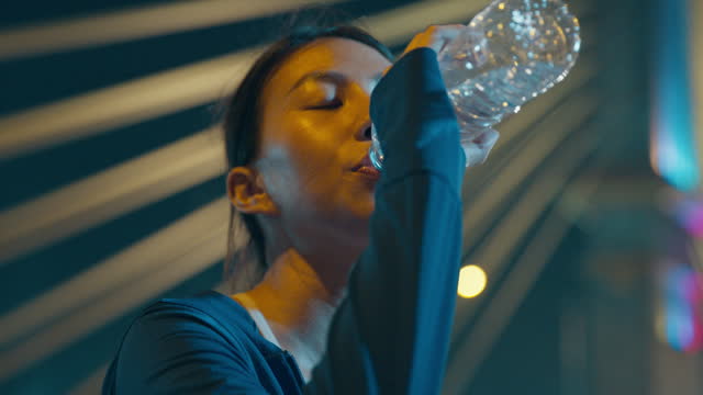 Closeup of happy young sporty Asian woman wear sports outfits drinking water from bottle through the city streets over bridge at night. Running club and exercise outdoor.