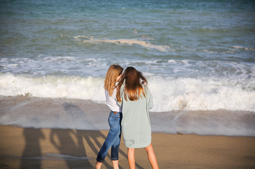 Two girls are standing on the beach on sunny day. Stylish hipsters near the waves on the sea. Women talking. Holiday travel concept. Barcelona Spain.