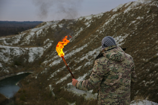 Soldier is holding torch with fire. Man in camouflage uniform and checkered bandana. Person in the mountains gorge. White hills and dry grass in the desert.