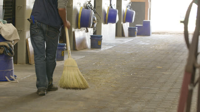 Man Using a Wicker Broom to Sweep the Alley of a Horse Barn