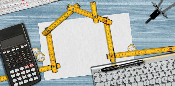 Folding wooden ruler in the shape of a house on a desk with blank sheet of paper with copy space, calculator, folding ruler, drawing compass, Euro coins, pencil and a computer keyboard.