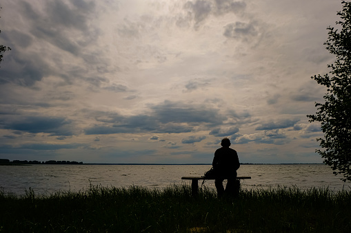 Person silhouette. The man 60 years is sitting on a wooden bench. Svitiaz Lake, Shatsk National Natural Park, Ukraine. Tranquil scene. Rest season.