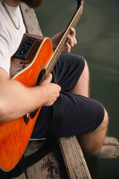 mężczyzna gra na gitarze, siedząc na drewnianym molo w pobliżu jeziora. - playing an instrument vertical blurred motion outdoors zdjęcia i obrazy z banku zdjęć