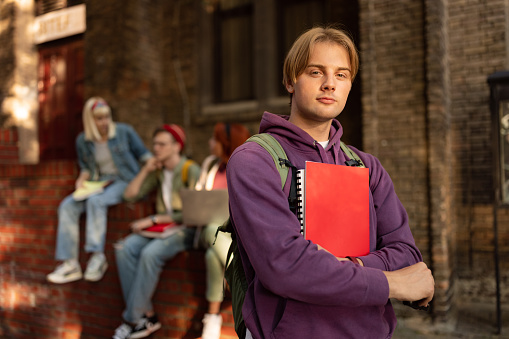 Young university student holding books in front of school building and looking at camera. His friends are in the background.