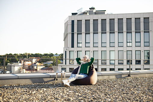 Wide view of relaxed person sitting in brown pouf chair with arms folded behind head against urban background. Attractive muslim woman in traditional clothing lazing on panoramic terrace in spring.