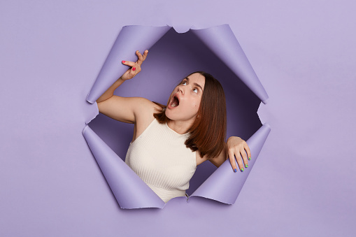 Shocked young brunette woman in casual clothing breaking through purple paper hole looking up with widely open mouth sees something amazement.