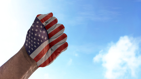 American flag in a clenched fist against a slightly cloudy blue sky background. Symbolizes the national spirit. Hands clenched