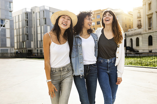 Cheerful brunette millennial female looking at camera standing in city street. Diverse female friends having fun together on summer vacation. Youth lifestyle and friendship concept.