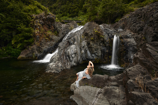 Young smiling woman relaxing on a rock with her eyes closed by the waterfall in nature.