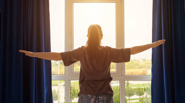 Brown-haired woman starts morning routine from exercises Brown-haired woman wearing t-shirt starts morning routine from simple exercises. Lady doing exercises looks at cityscape outside window, sunlight vista posterior stock pictures, royalty-free photos & images