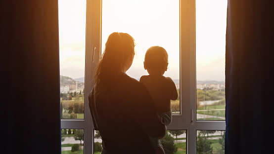 Adult woman with toddler girl in arms opens curtains together on panoramic windows. Mother and daughter look at morning cityscape from home