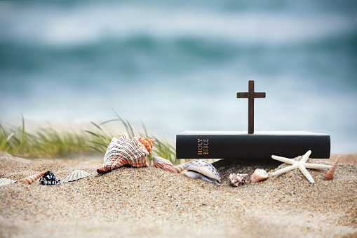 Church summer retreat concept with bible book, cross of Jesus Christ and shells, conch and starfish on sandy beach and sea with waves Summer bible school summer camp background